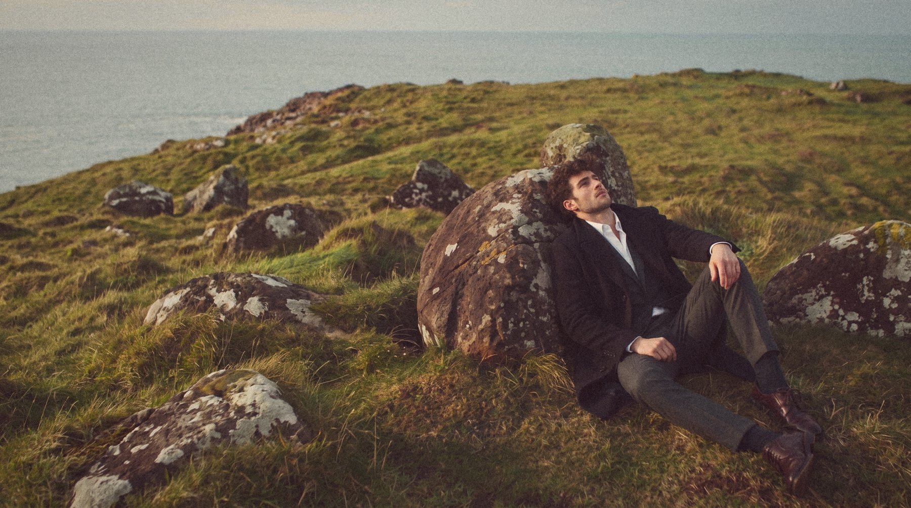 A man in a black overcoat and grey suit leans back against a boulder on a clifftop in low sun.