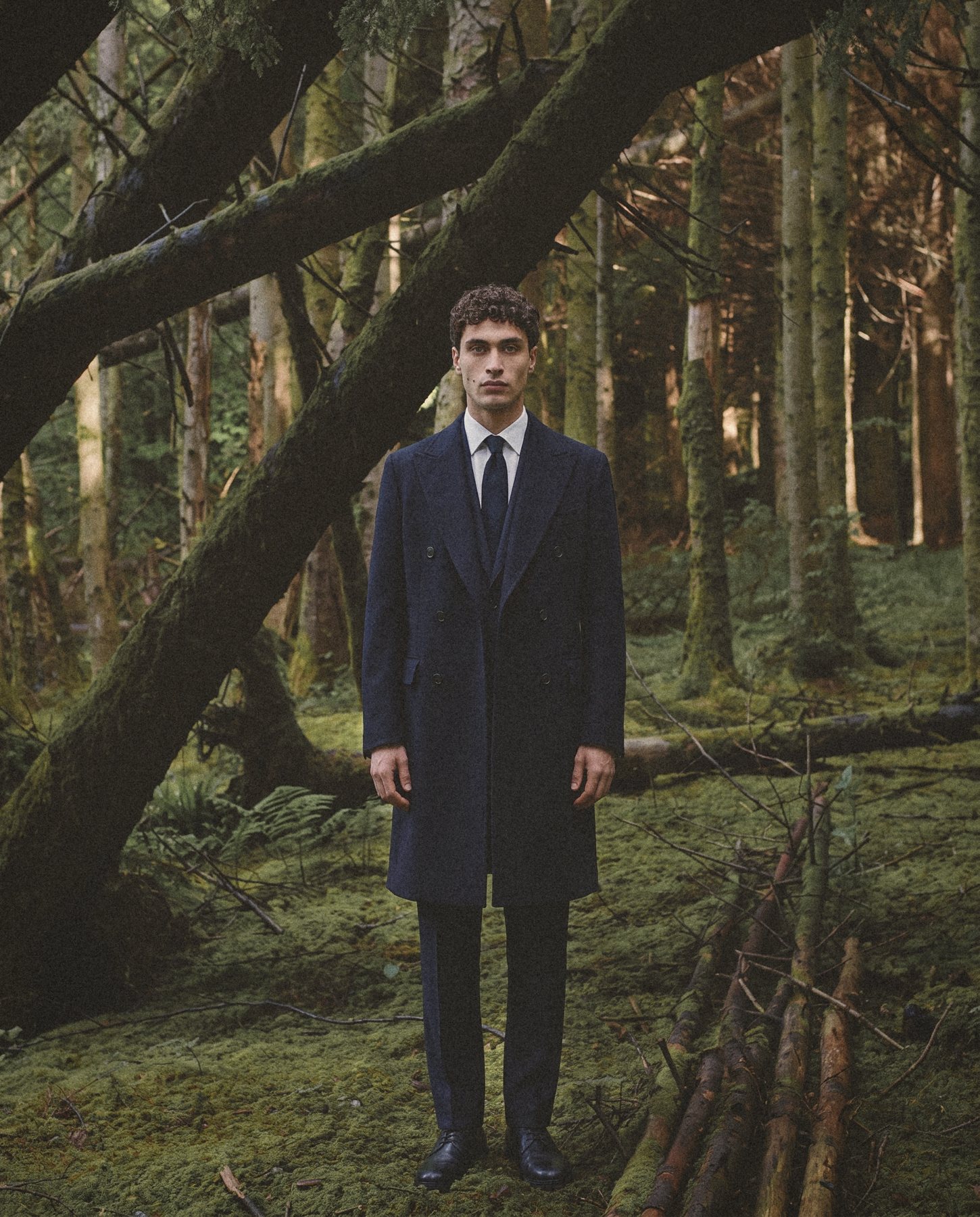 A man in a navy double breasted overcoat stands in a mossy forest.