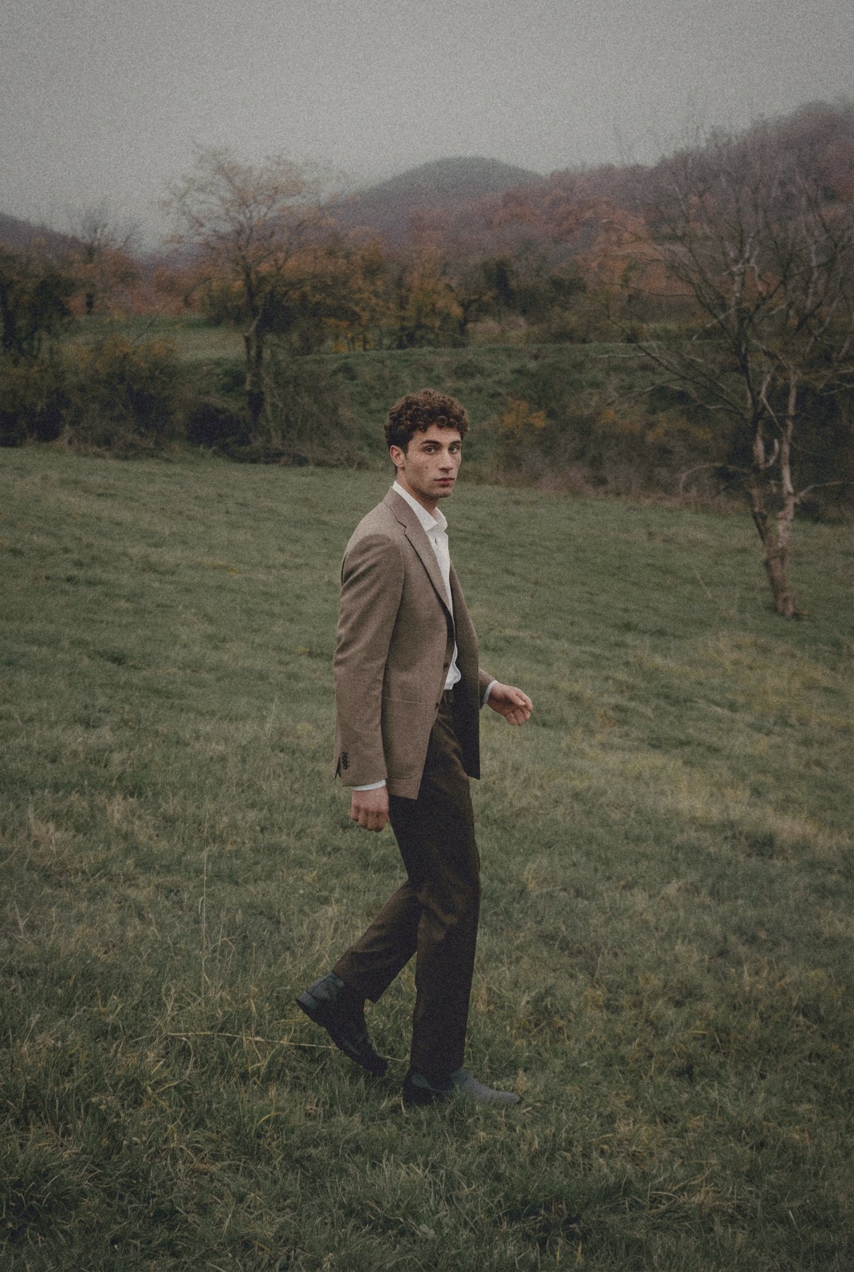 A man in a taupe jacket walks across a foggy meadow and looks back.