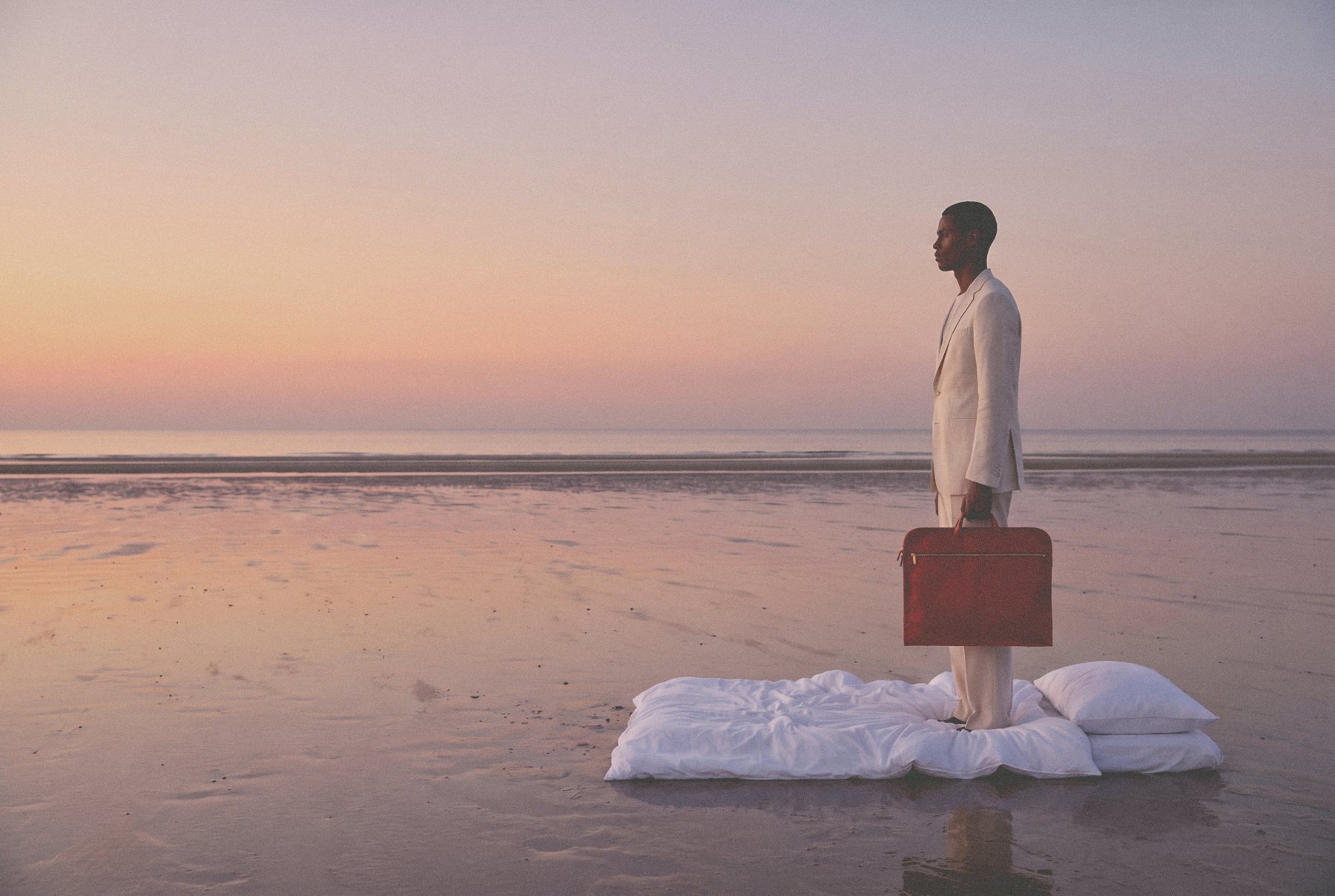 A man in a cream suit holding a red leather briefcase stands on a white bed laid out on a wet beach at dusk.