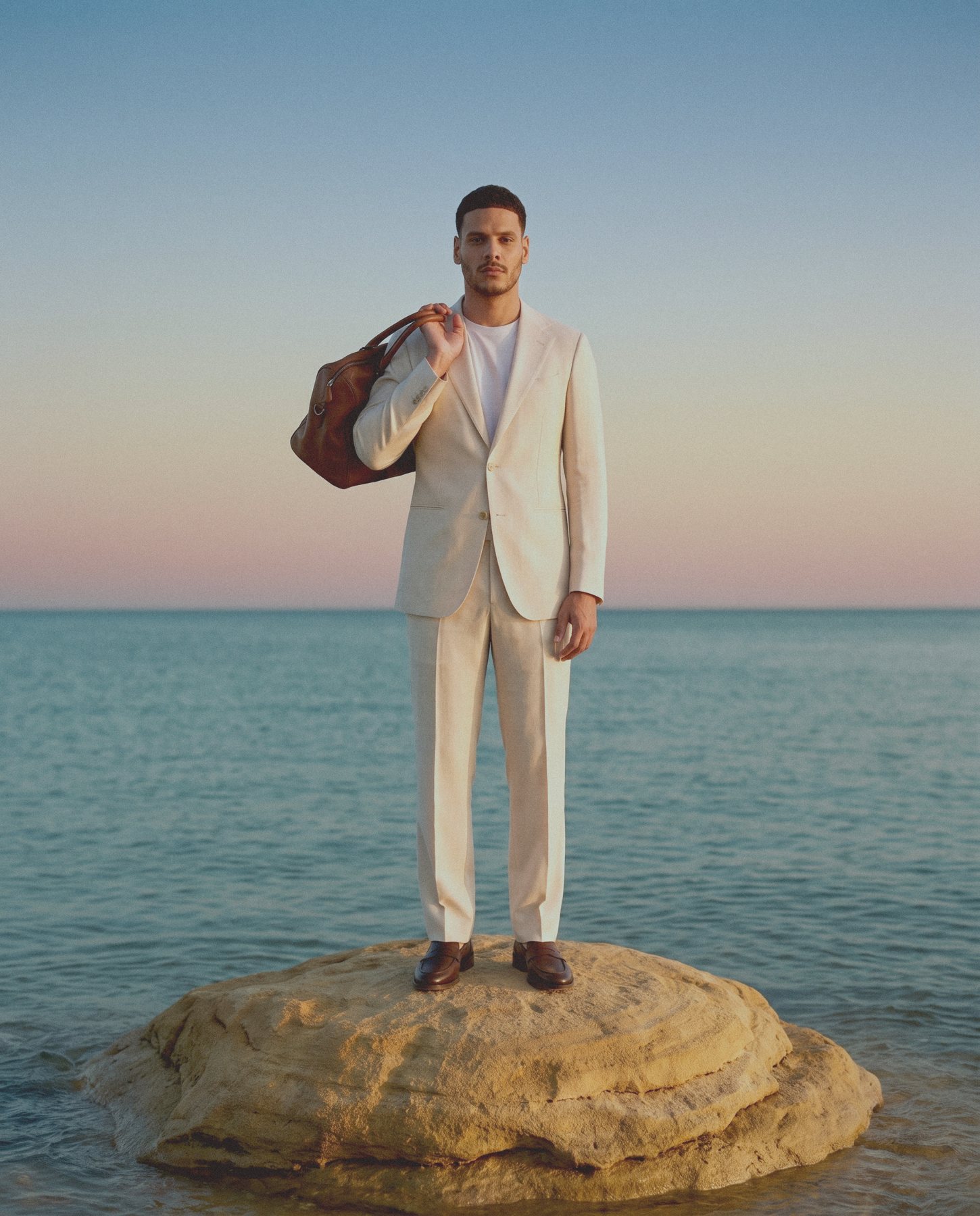 A man in a cream suit with a brown leather holdall over his shoulder stands on a rock in the sea at dusk.
