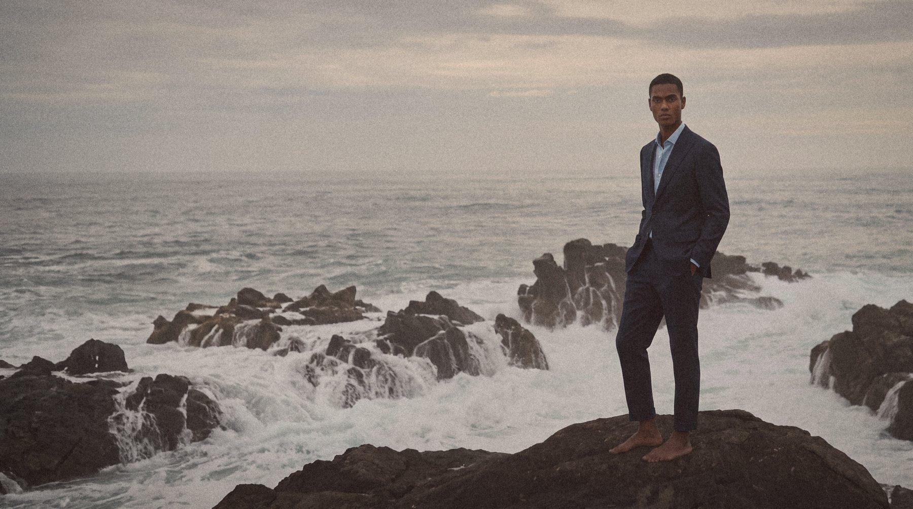 A man in a navy suit stands on dark rocks as the surf breaks behind him.