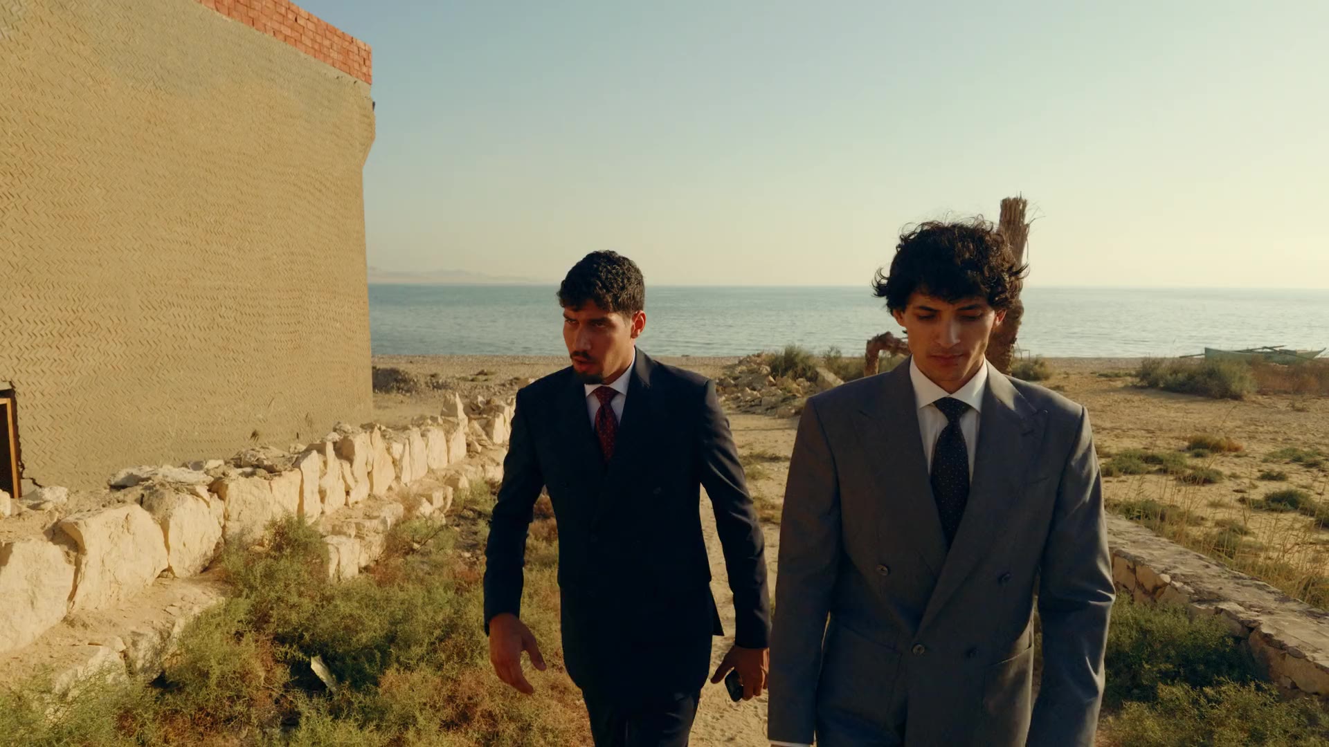 Two young men in double breasted suits, one navy and one grey, walk towards the camera along a stone wall by the lake in the morning sun.