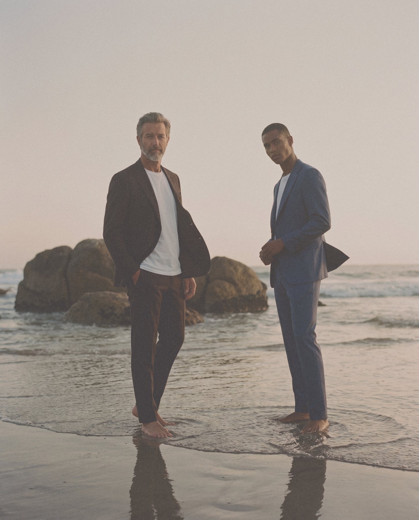 Two men, one grey bearded in a dark brown jacket and one in a blue suit, stand barefoot at the water’s edge at sunset.