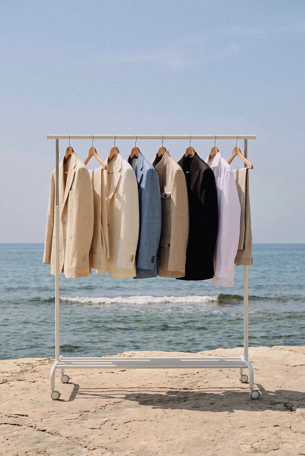 A clothes rail of linen and wool jackets stands on a rock above the sea.