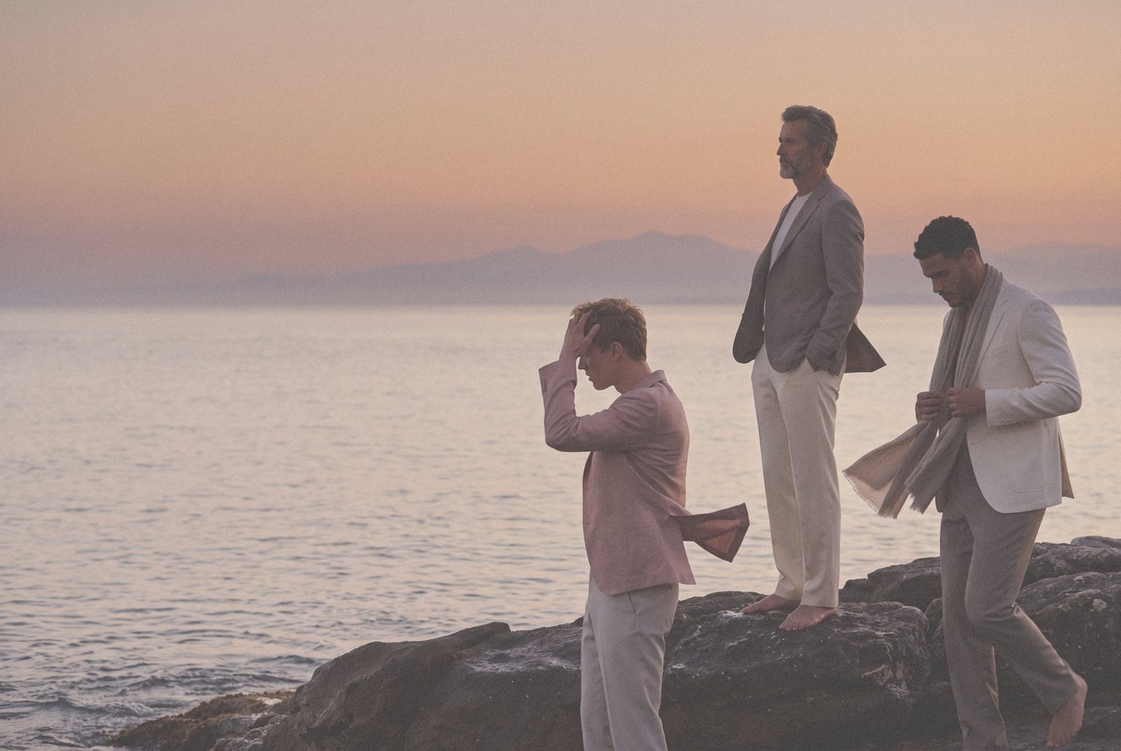 Three men in light tailoring stand on rocks by the sea at dusk.