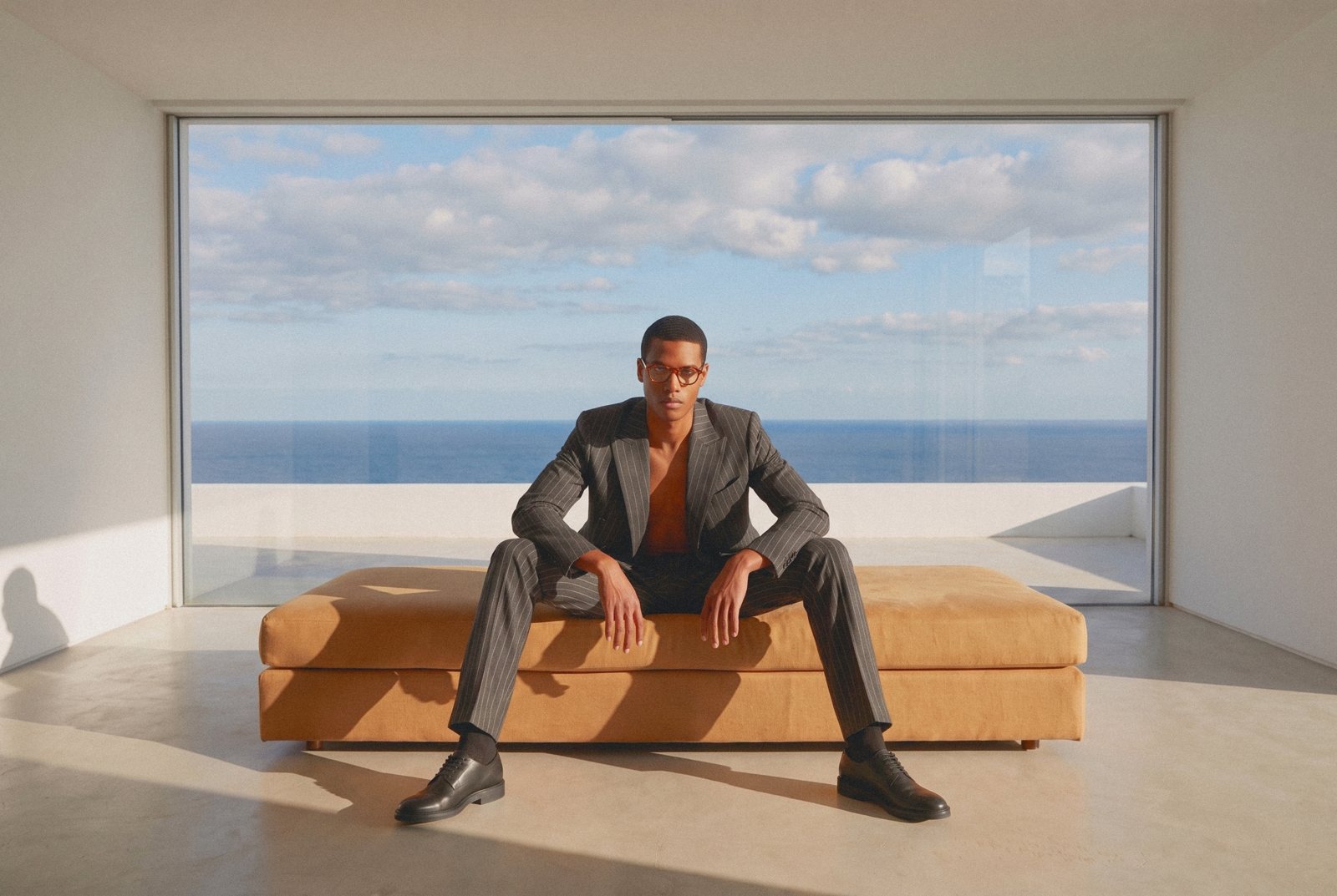 A man in a grey suit sits on a leather bench before a wide window over the sea.