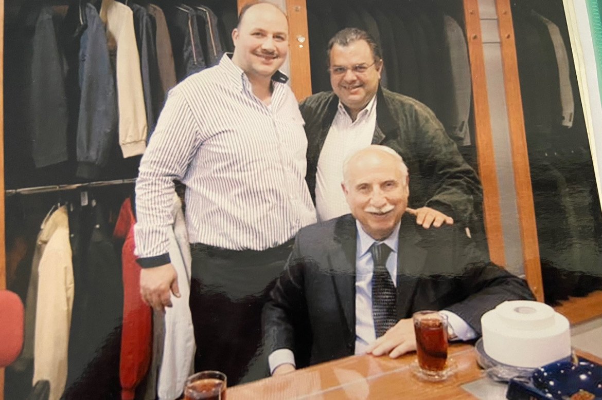 Mohamad Hajjo seated at a table in the shop with his son Omar Hajjo and another man, coats on the rails behind them.